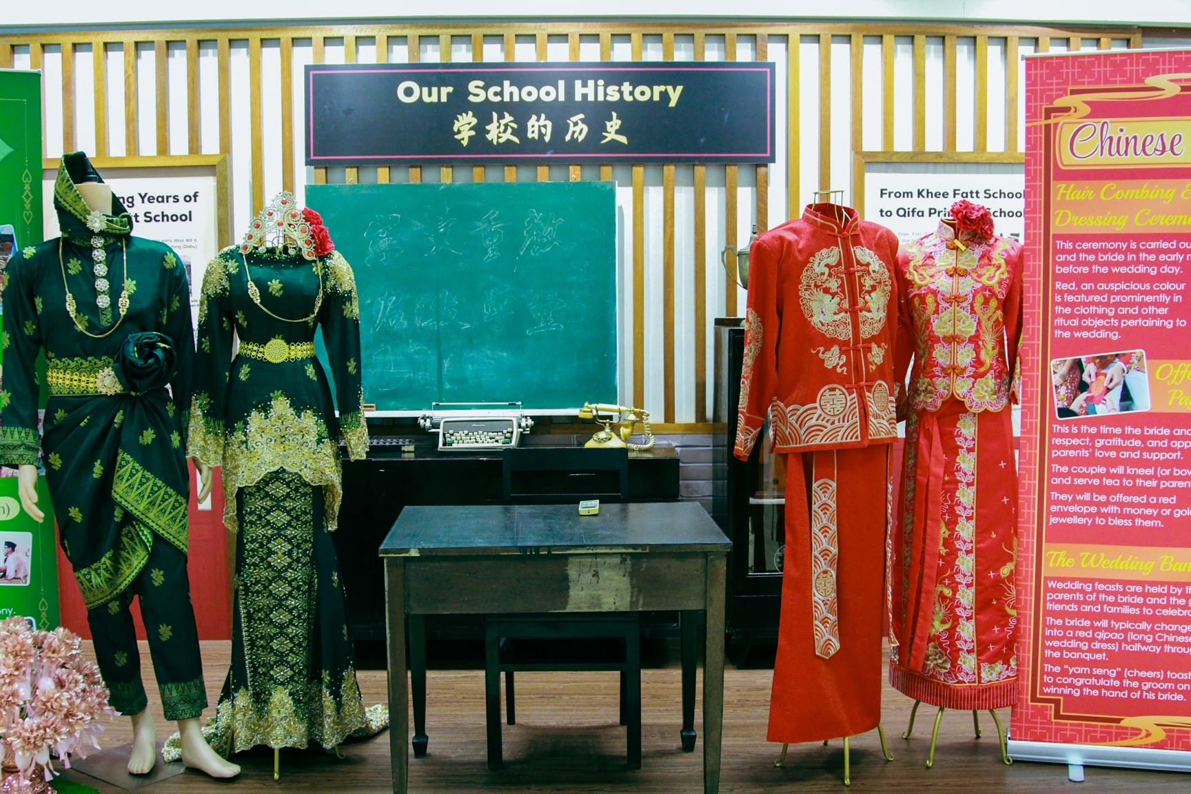 two mannequins in traditional Malay wedding attire in dark green with gold embroidery on the left, and a red Chinese wedding outfit (xiuhe fu) with gold dragon and phoenix embroidery on the right. In the centre, a vintage chalkboard, typewriter, and rotary telephone are arranged around an old wooden desk. A red pull-up banner on the far right describes Chinese wedding customs.
